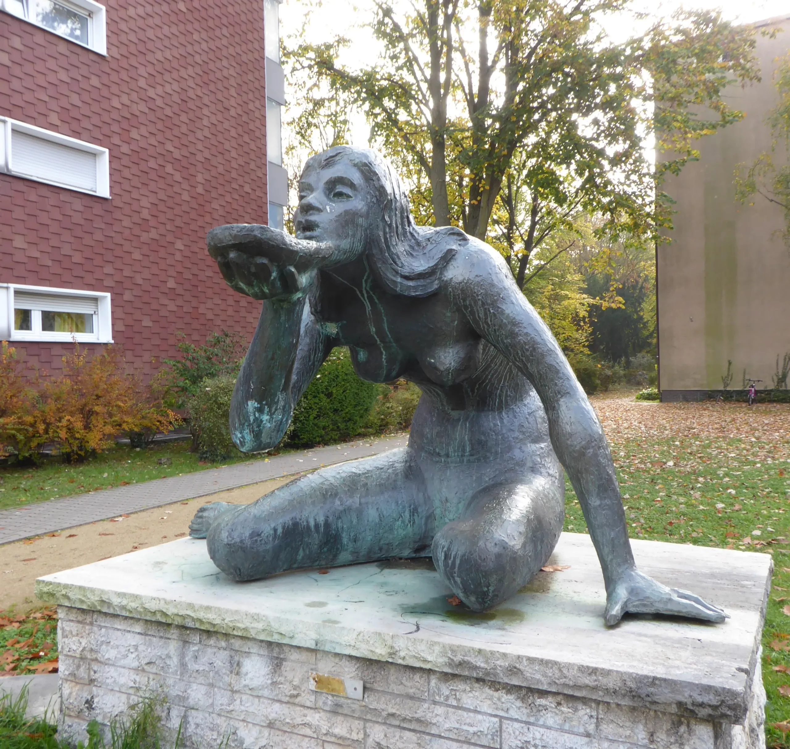 Bronzestatue einer knienden Frau, die eine Muschel an die Lippen hält, als wolle sie daraus trinken; sie steht auf einem Steinsockel im Freien in der Nähe von Gebäuden und Bäumen.