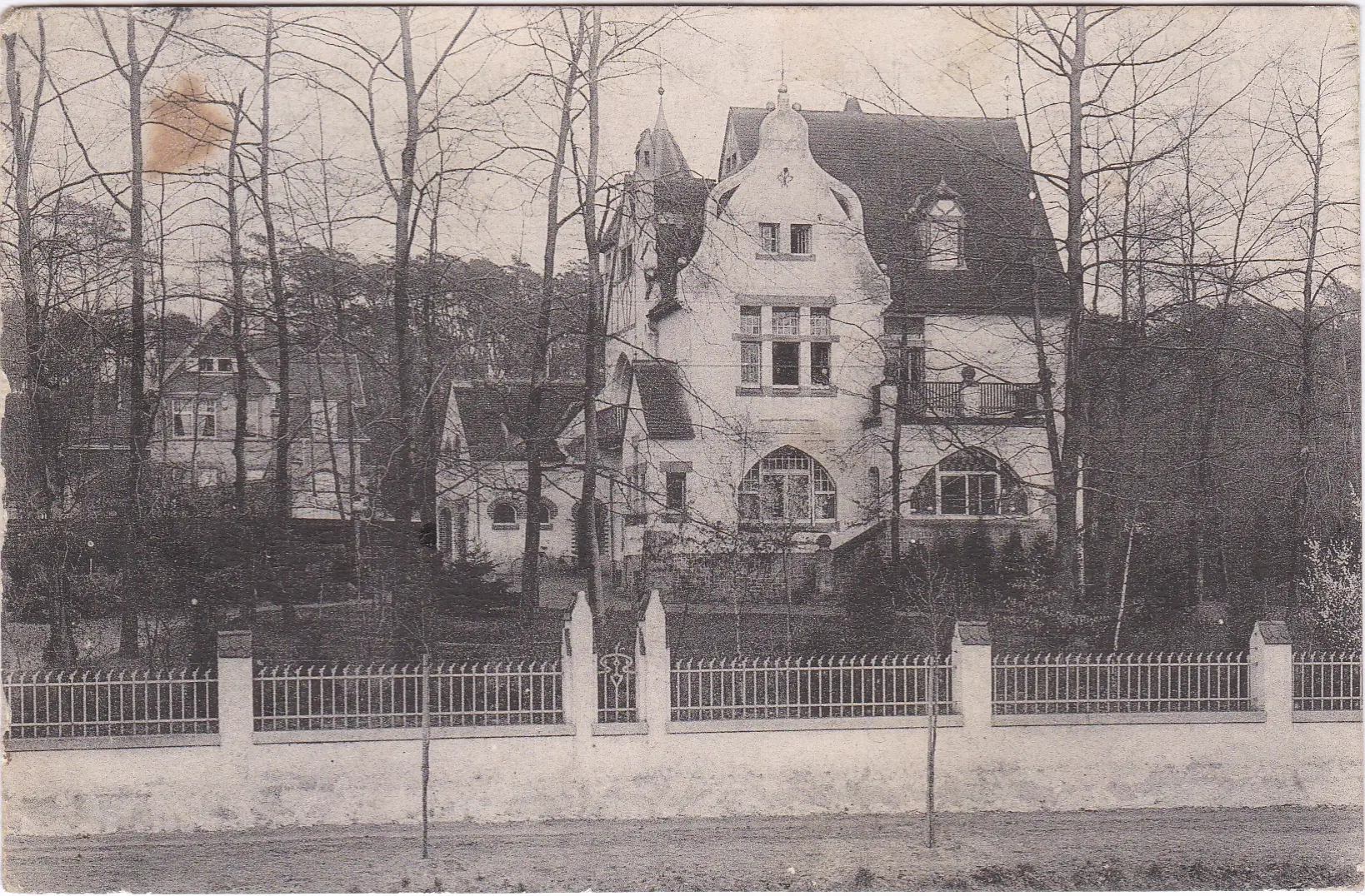Ein altes Schwarz-Weiß-Foto eines großen, prunkvollen Hauses mit Bogenfenstern und steilen Dachlinien, umgeben von kahlen Bäumen und eingefasst von einem verzierten Zaun. Im Hintergrund ist teilweise ein weiteres Haus zu sehen.