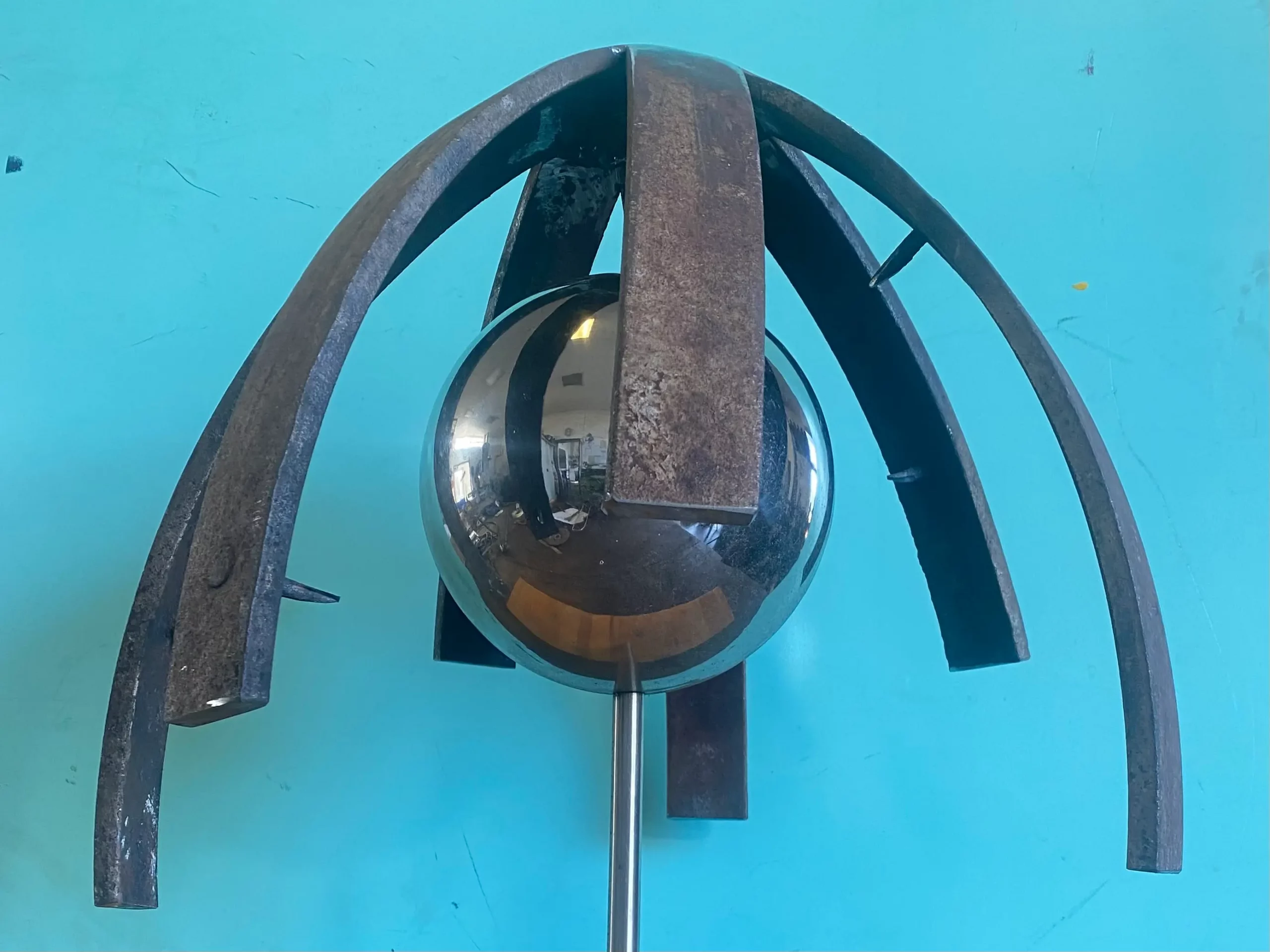 Eine Metallskulptur mit einer reflektierenden Kugel, die von vier geschwungenen, verrosteten Metallstangen umschlossen wird, vor einem hellblauen Hintergrund.