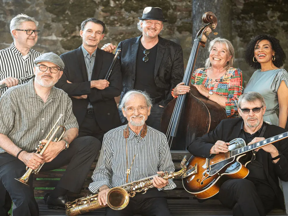 Acht lächelnde Erwachsene posieren gemeinsam mit Musikinstrumenten – darunter eine Trompete, ein Saxophon, eine Gitarre und ein Kontrabass – vor einer rustikalen Steinmauer. Einige stehen, andere sitzen davor; alle sind leger bis halbformell gekleidet.