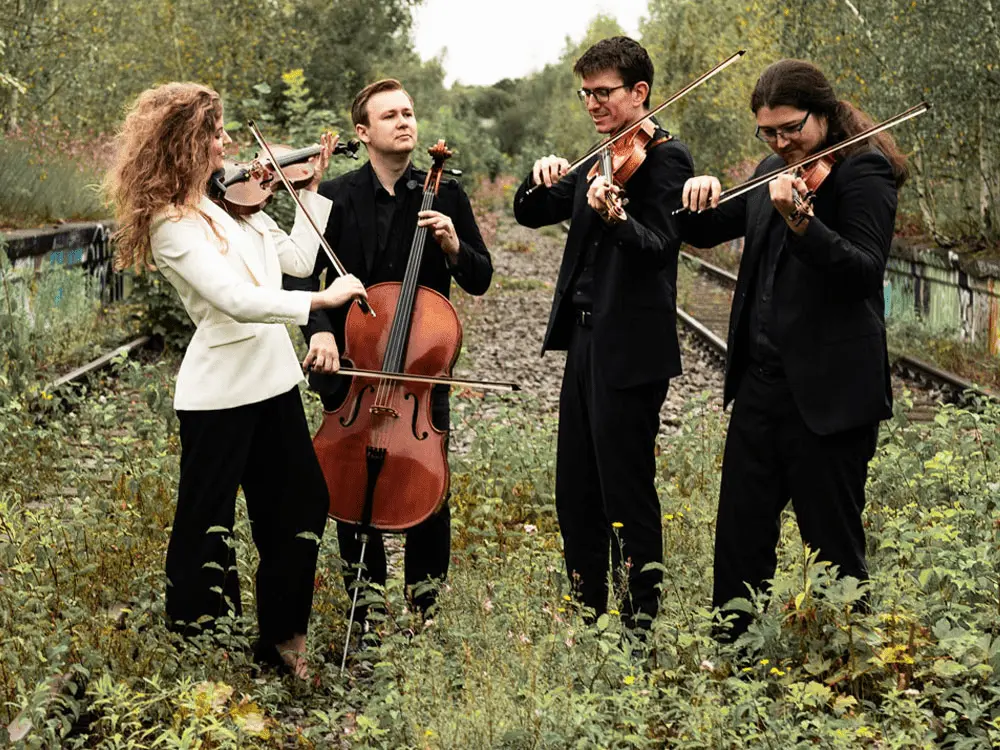 Vier Musiker, die Streichinstrumente spielen – zwei Violinen, eine Bratsche und ein Cello –, stehen im Freien auf überwucherten Bahngleisen; sie tragen schwarze Abendgarderobe, bis auf einen, der einen weißen Blazer trägt.