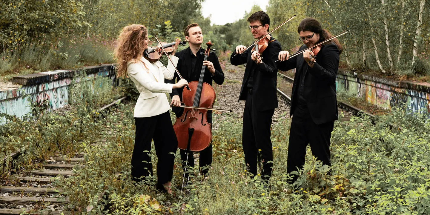 Vier Musiker in Abendgarderobe spielen Streichinstrumente auf überwucherten, mit Graffiti übersäten Bahngleisen, die von Bäumen umgeben sind. Zu der Gruppe gehören zwei Geiger, ein Bratschist und ein Cellist.