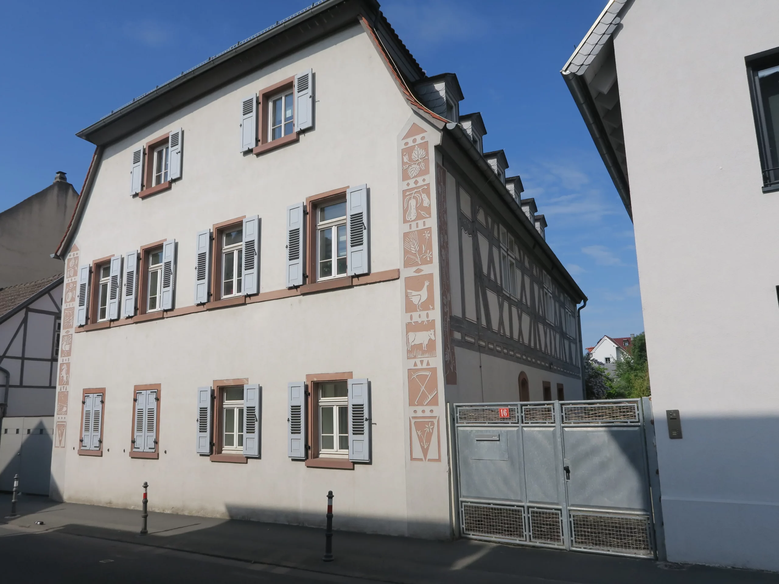 Ein weißes, dreistöckiges Gebäude mit bemalten Fensterläden und Verzierungen an der Ecke steht in einer ruhigen Straße, neben einem Metalltor und einem weiteren weißen Gebäude unter strahlend blauem Himmel.