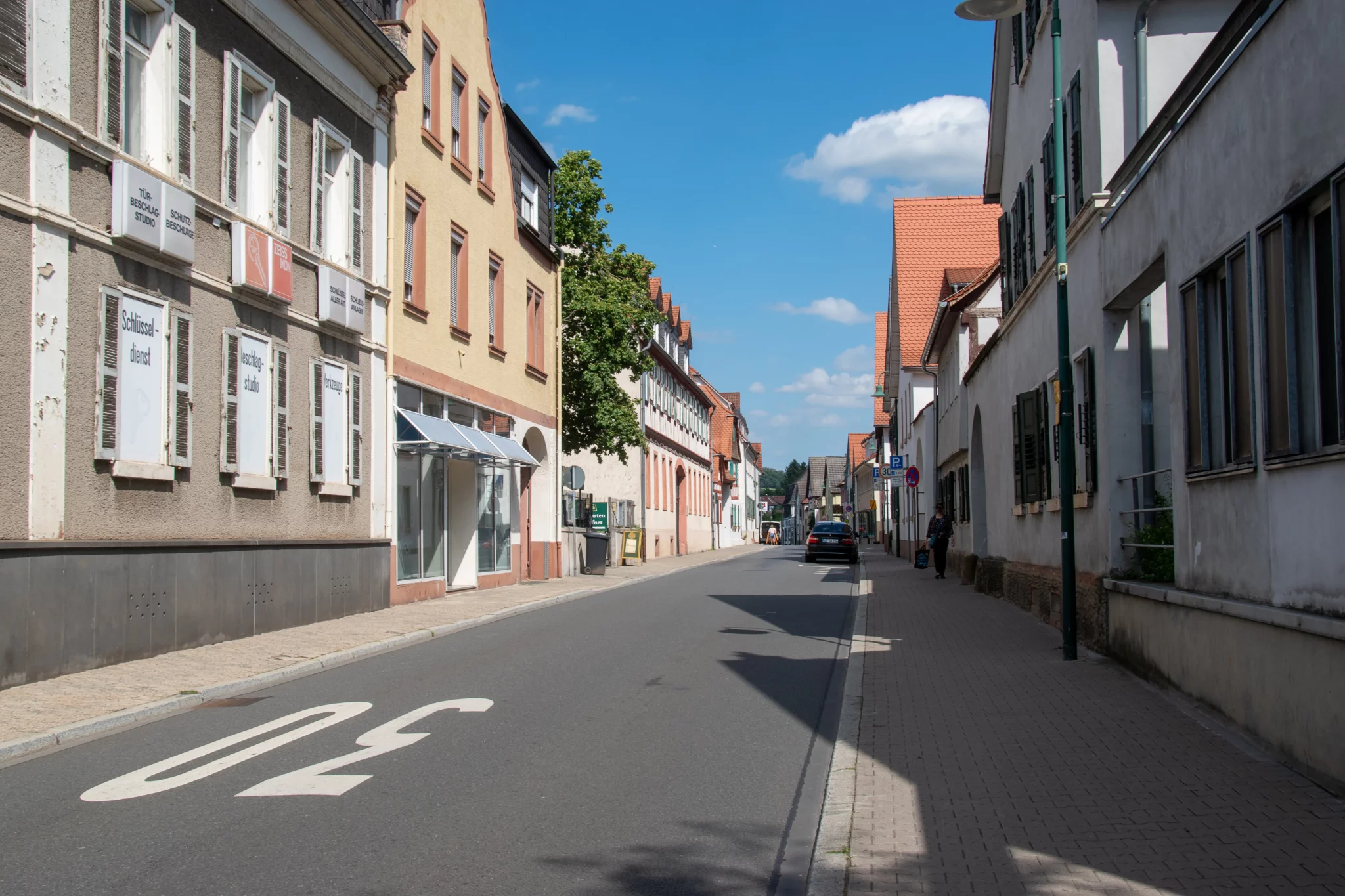 Eine ruhige, menschenleere Straße mit Häusern und Gebäuden auf beiden Seiten, einer auf der Fahrbahn aufgemalten Geschwindigkeitsbegrenzung von 30 km/h, geparkten Autos und einem strahlend blauen Himmel.