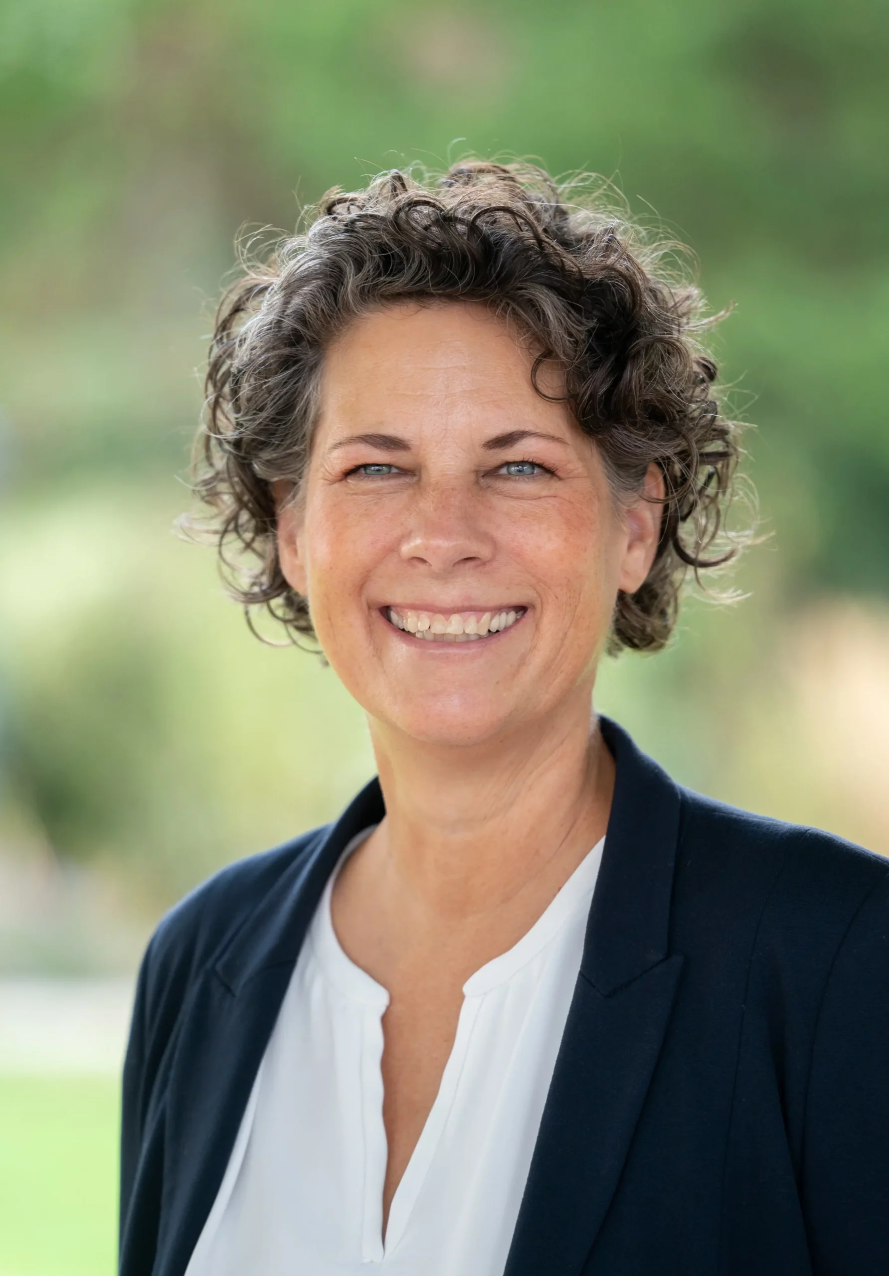 Eine Frau mit kurzen Locken lächelt in die Kamera. Sie trägt einen marineblauen Blazer und ein weißes Oberteil und steht im Freien vor einem verschwommenen grünen Hintergrund.