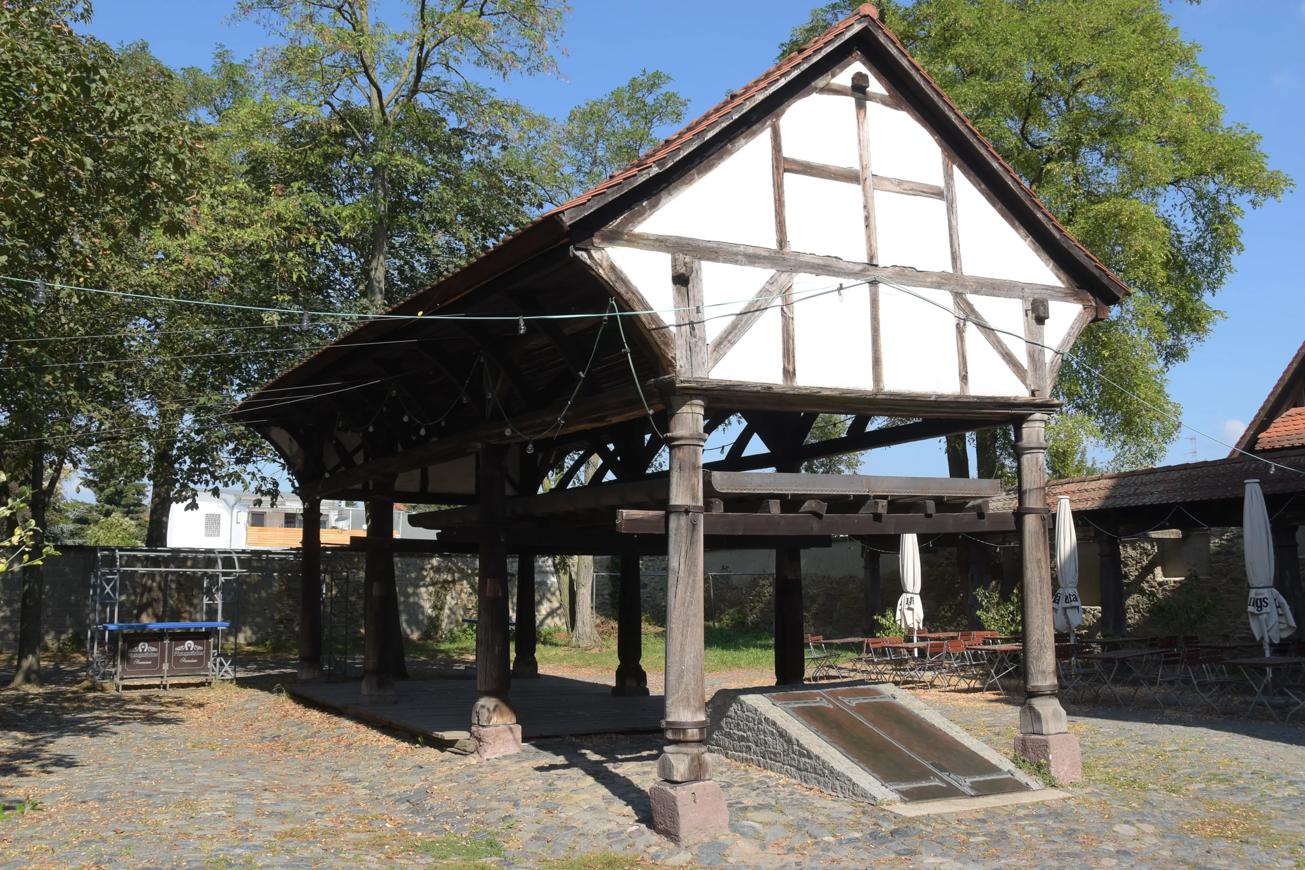 Ein historisches Waschhaus aus Holz mit einem geneigten Waschbereich aus Stein steht auf Steinsäulen in einem sonnigen Innenhof, umgeben von Bäumen und Kopfsteinpflaster.