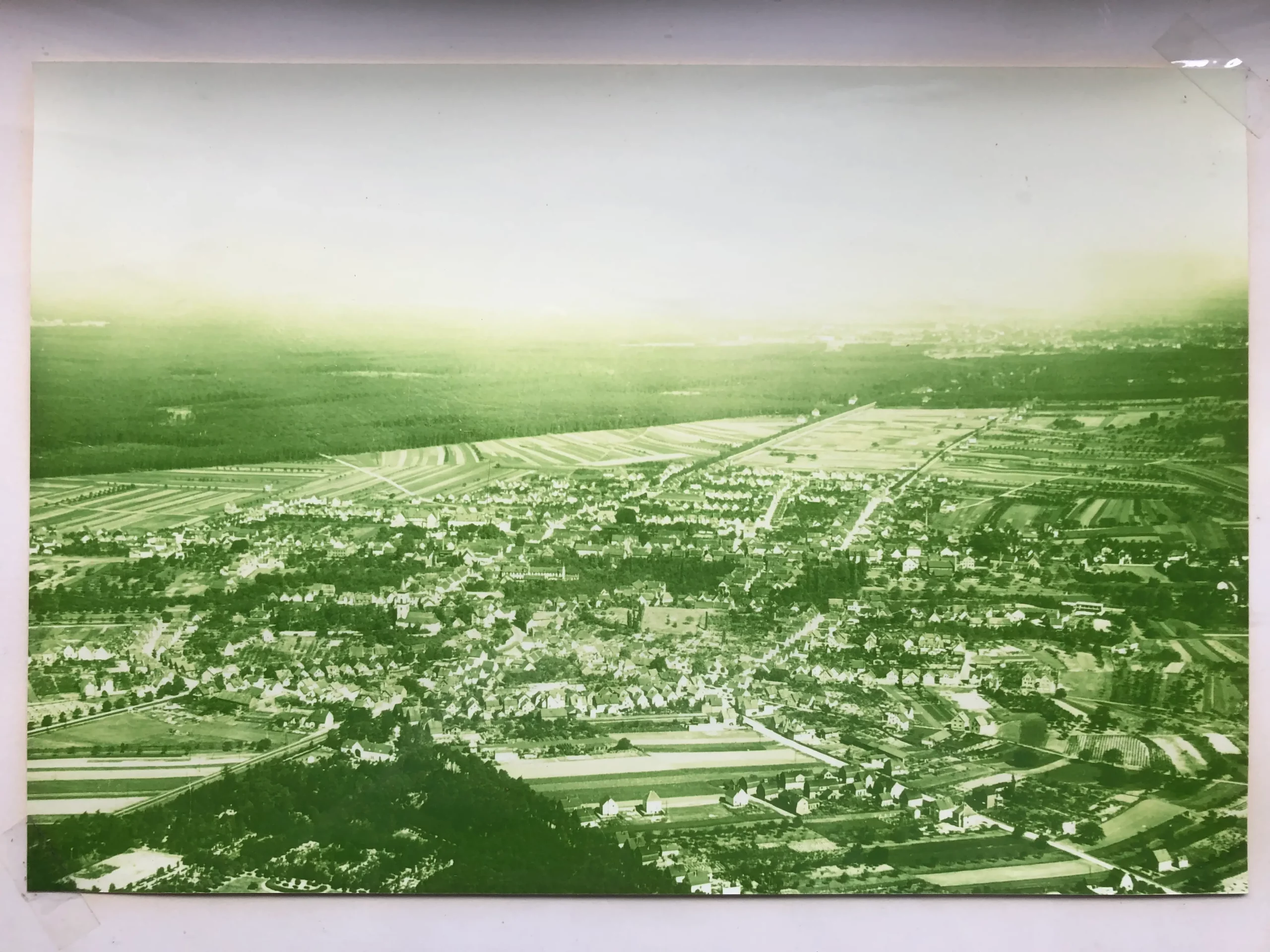 Luftaufnahme einer Stadt mit dicht beieinander stehenden Häusern, Straßen und Feldern, umgeben von Wald. Das Foto weist einen grünlichen Farbton und einen dunstigen Himmel auf.
