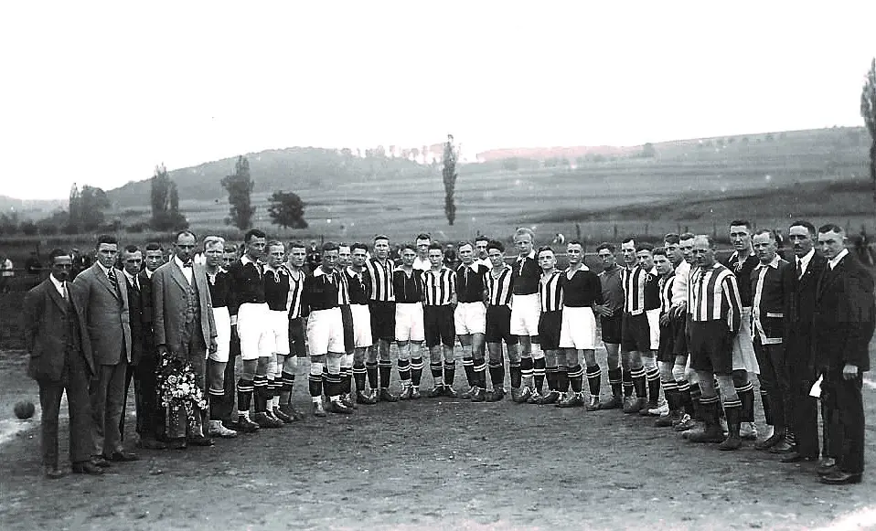Zwei Fußballmannschaften und mehrere Männer in Anzügen stehen gemeinsam in einem Halbkreis auf einem unbefestigten Spielfeld und posieren für ein Gruppenfoto vor dem Hintergrund von Hügeln und Bäumen. Die Spieler tragen gestreifte Trikots und Shorts.
