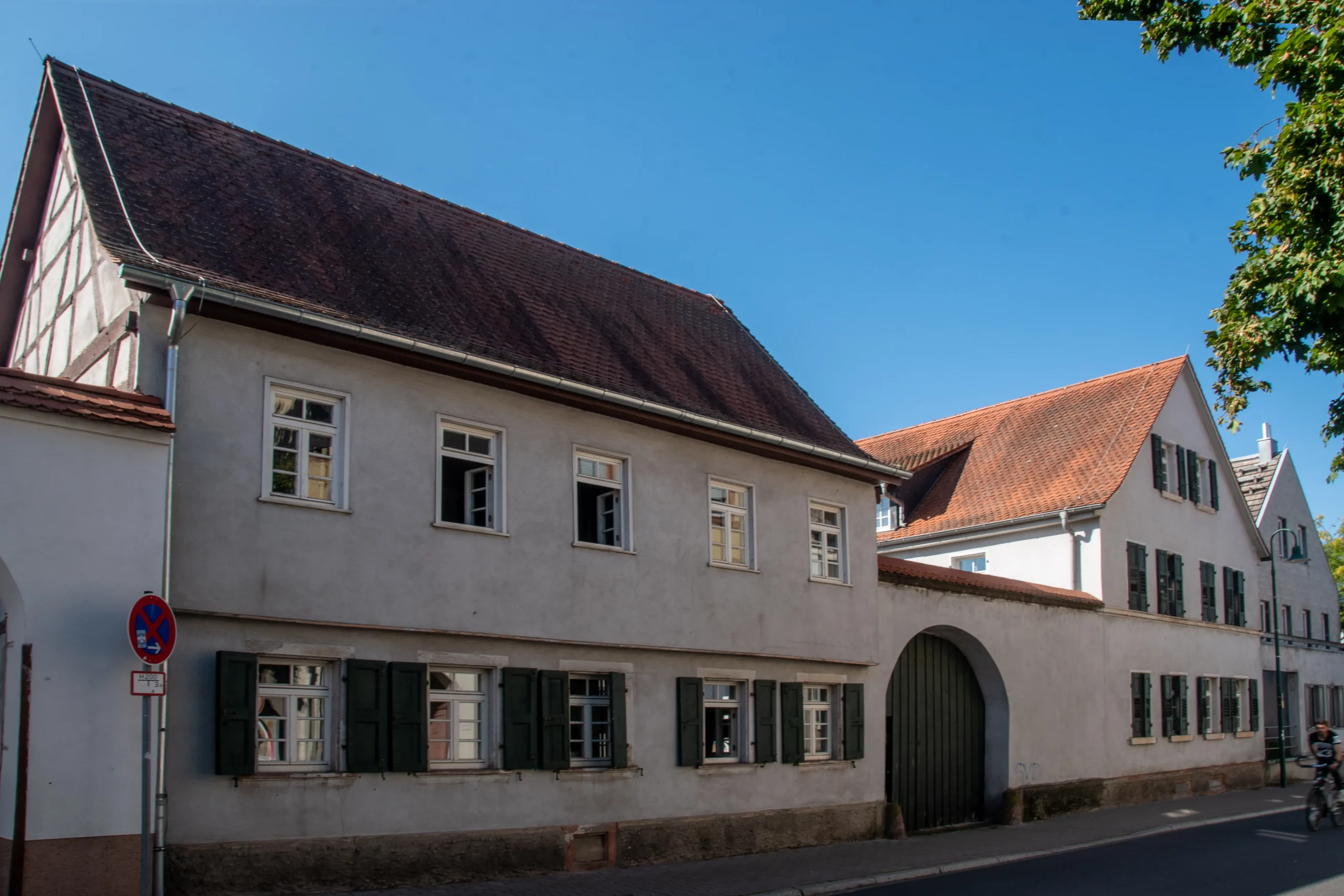 Ein Straßenansicht eines alten, beigen Gebäudes mit grünen Fensterläden und einem großen Torbogen, neben einem weiteren Gebäude mit roten Dachziegeln, unter einem strahlend blauen Himmel.