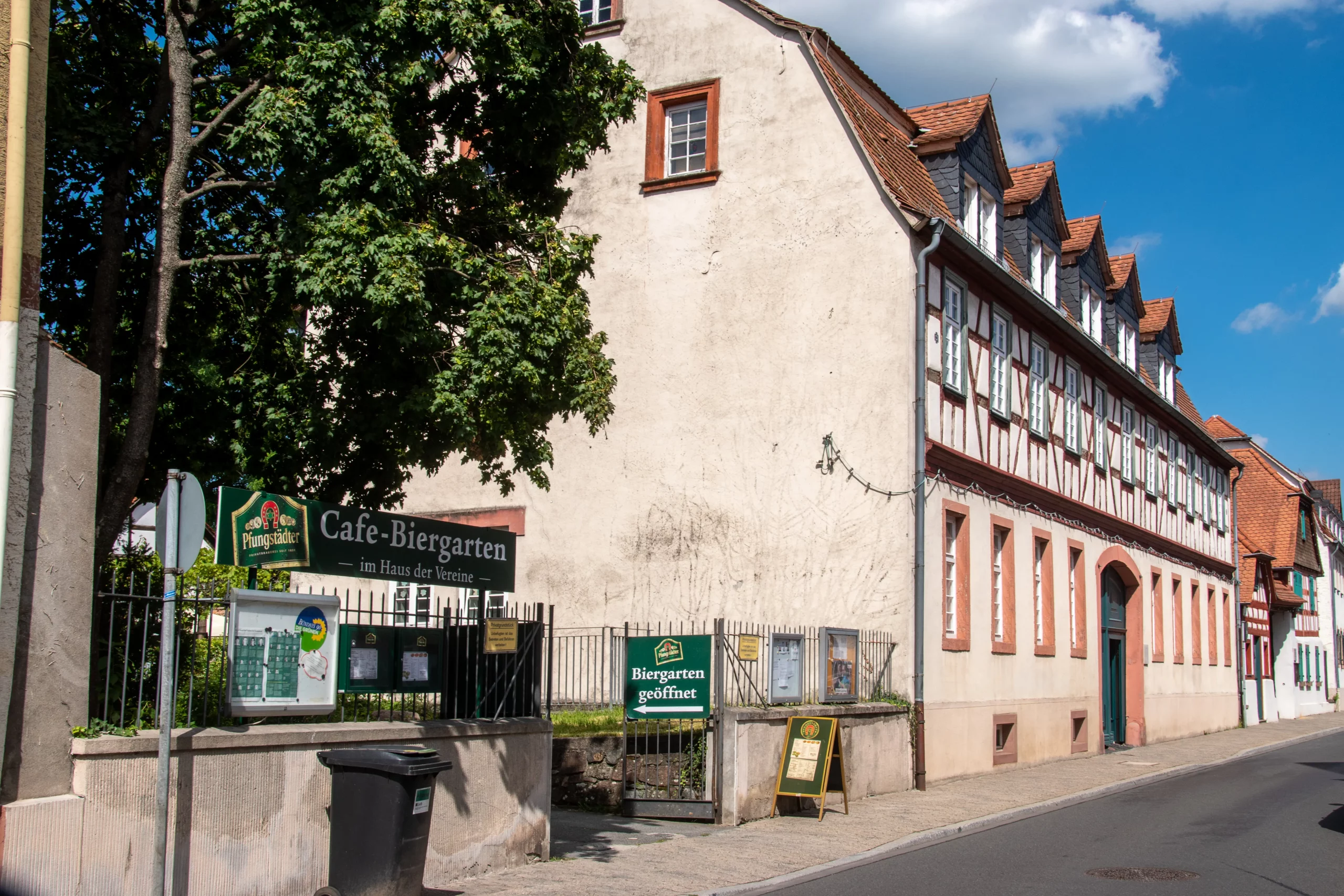 Eine Straßenansicht eines traditionellen deutschen Fachwerkhauses. Schilder weisen auf ein Café mit Biergarten und Sitzgelegenheiten im Freien hin. Im Hintergrund sind Bäume und ein klarer Himmel zu sehen.