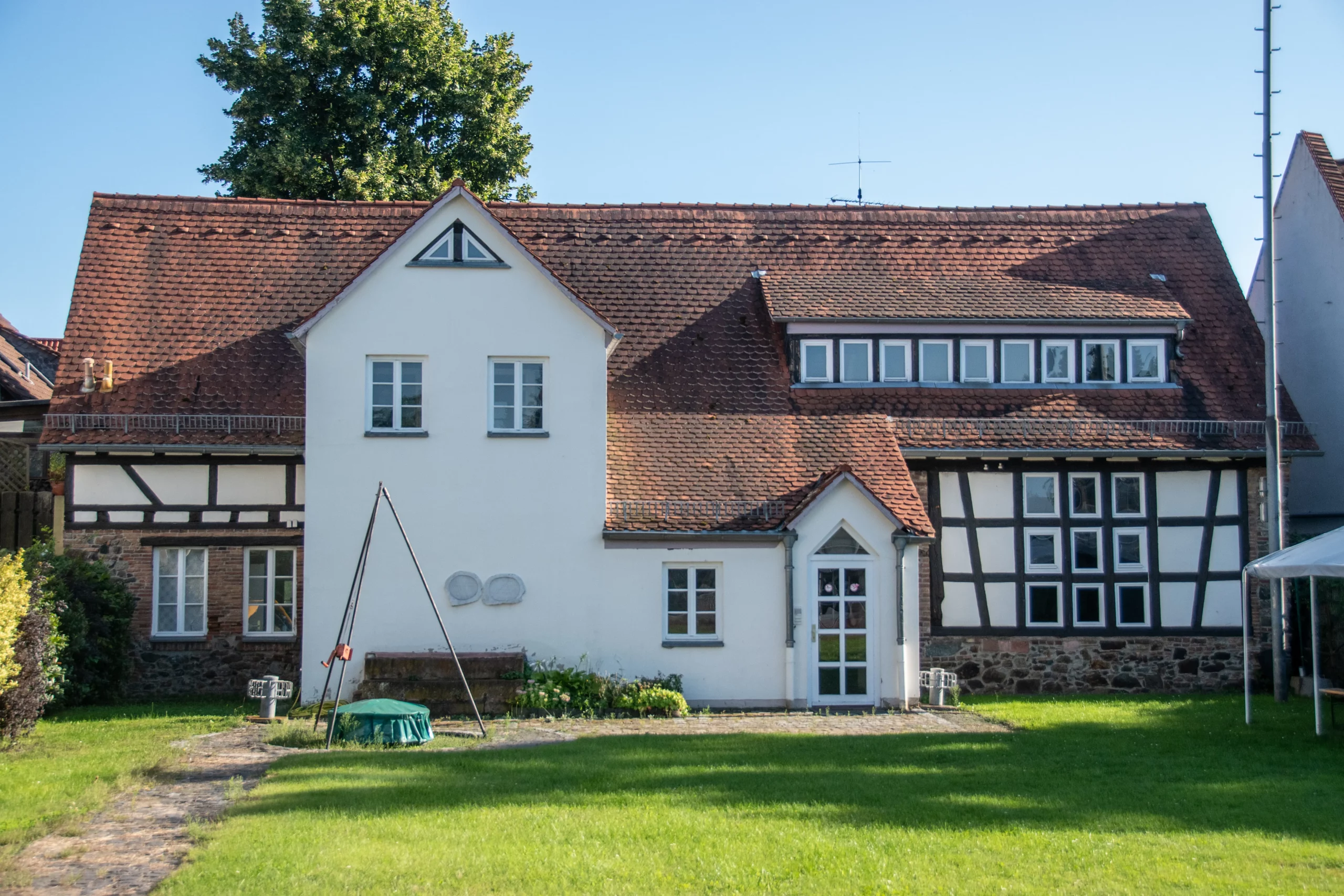 Ein traditionelles europäisches Haus mit weißen Wänden, sichtbaren Holzbalken, einem roten Ziegeldach und einer Schaukel im grasbewachsenen Garten an einem sonnigen Tag.