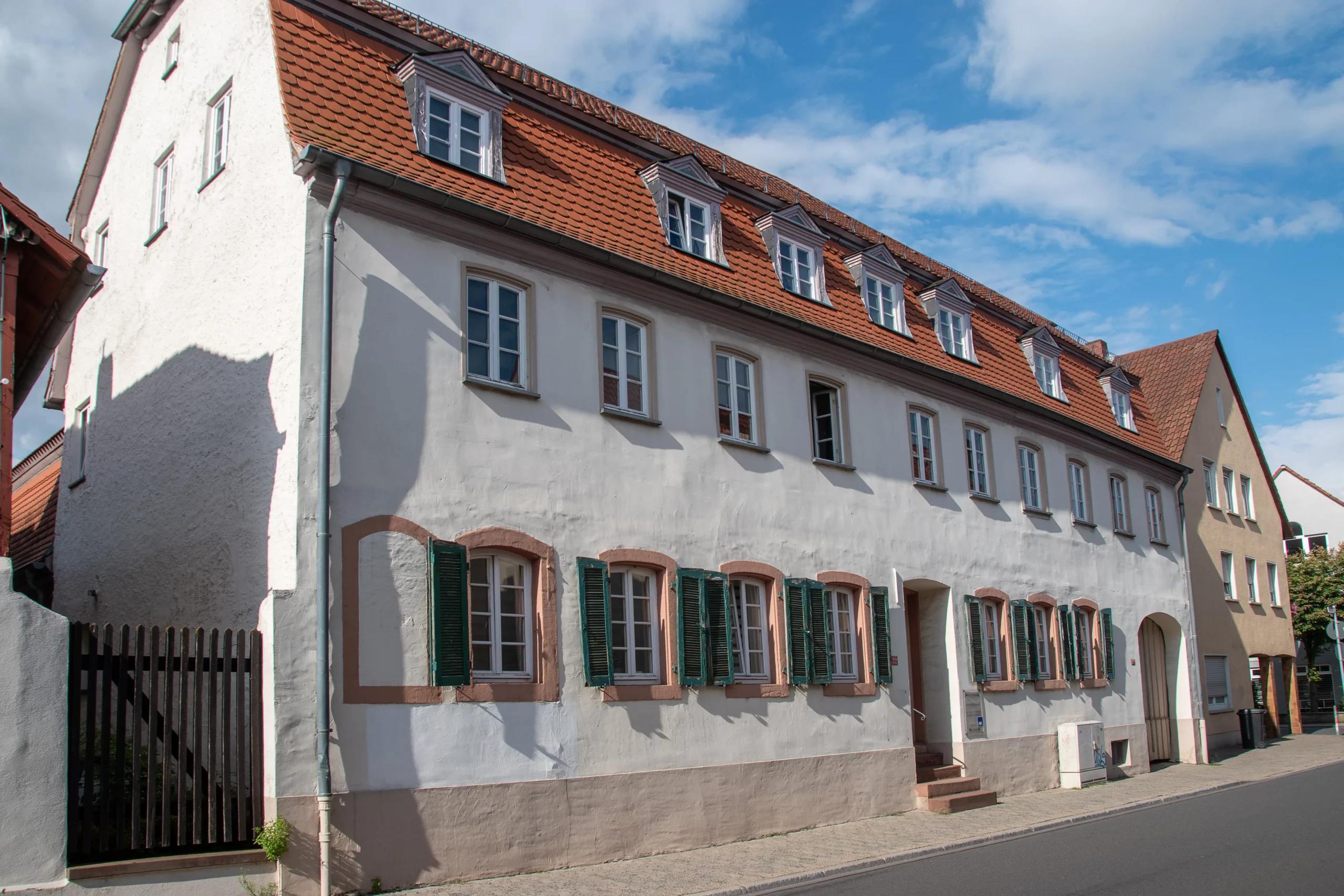 Ein großes, weißes, dreistöckiges Gebäude mit einem roten Ziegeldach und vielen Fenstern steht an einer ruhigen Straße unter einem blauen Himmel mit Wolken.