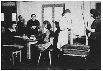 Schwarz-Weiß-Foto von Frauen und Mädchen, die in einem Klassenzimmer oder einer Werkstatt an Tischen arbeiten; einige sitzen, andere stehen und sind mit verschiedenen Aufgaben beschäftigt.