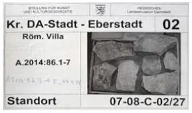 Etikett für archäologische Fundstücke aus Eberstadt, Deutschland, mit Referenznummern, der Angabe „Röm. Villa“, einem Standortcode und einem Foto von Steinfragmenten auf der rechten Seite.