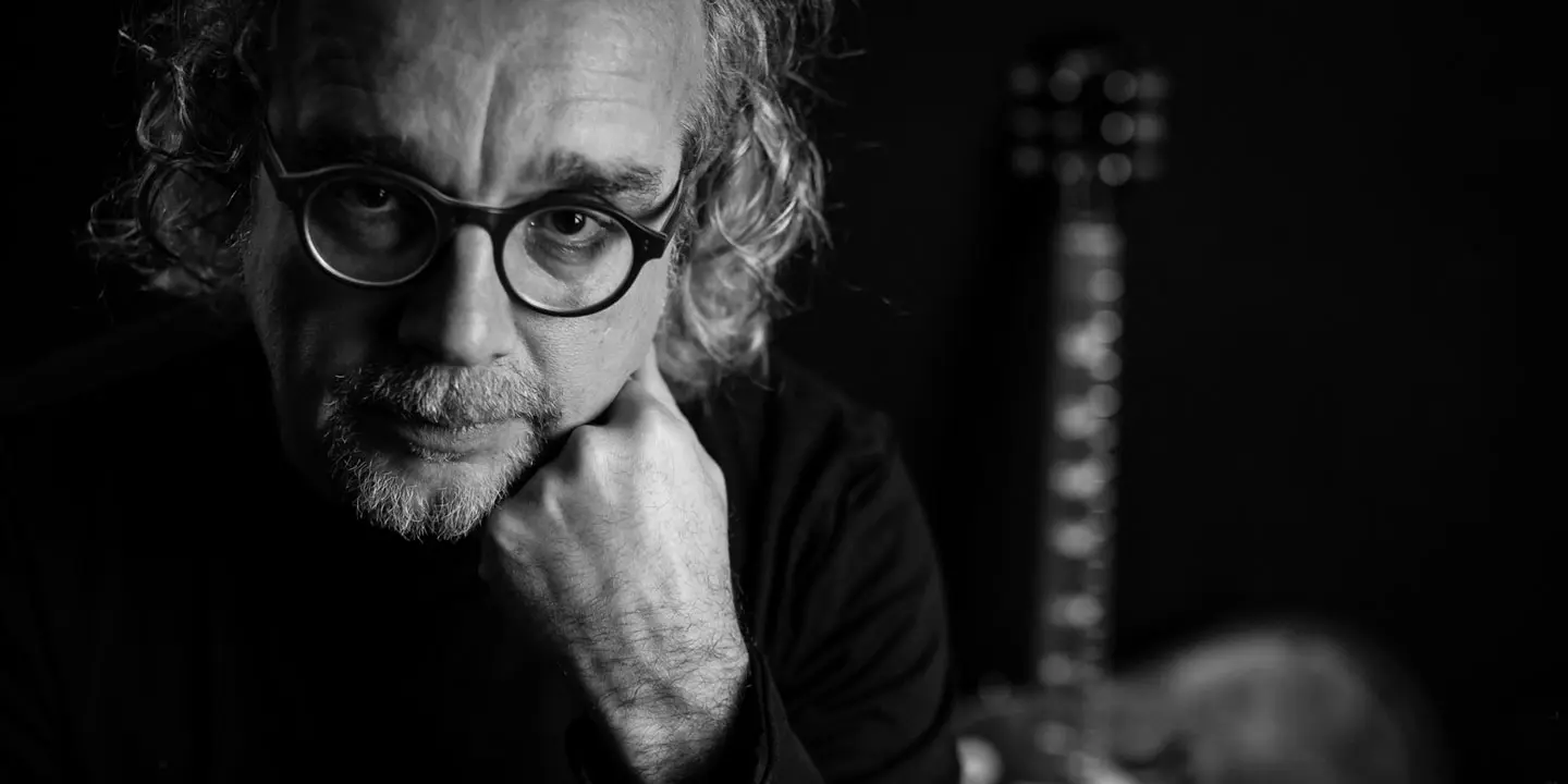 Schwarz-Weiß-Foto eines Mannes mit lockigem Haar und Brille, der sein Kinn auf die Hand stützt und in die Kamera blickt. Im Hintergrund ist eine verschwommene Gitarre zu sehen.