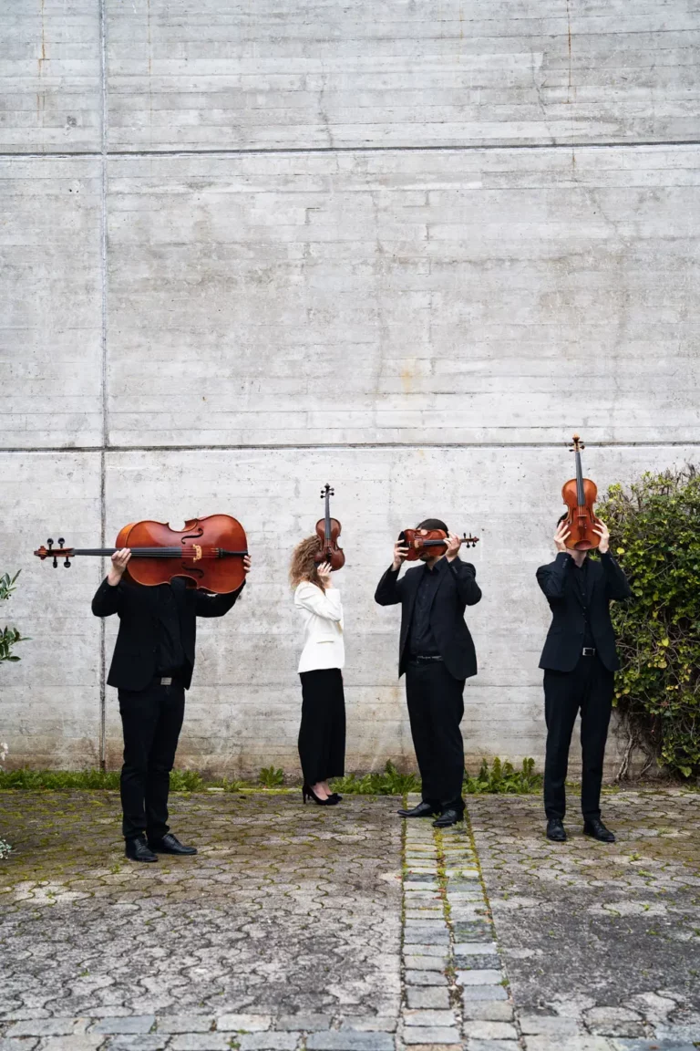 Vier Personen in schwarzen Anzügen und eine in einer weißen Jacke stehen vor einer Betonwand und verbergen ihre Gesichter hinter Streichinstrumenten, darunter ein Cello, eine Bratsche und Geigen.