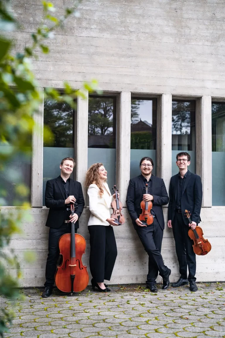 Vier Musiker in Abendgarderobe stehen draußen vor einem Betongebäude mit hohen Fenstern und halten jeweils ein Streichinstrument in der Hand: zwei Geigen, eine Bratsche und ein Cello. Die Gruppe wirkt lächelnd und entspannt.