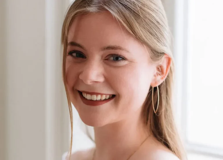 Eine junge Frau mit langen blonden Haaren lächelt in die Kamera. Sie trägt Creolen und ist dezent geschminkt. Sie steht vor einem sanft beleuchteten Fenster vor einem hellen Hintergrund, was an die zeitlose Eleganz von Schumann und Brahms erinnert.